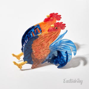 May include: A pair of rooster-shaped brooches. The brooches feature a vibrant color palette of red, orange, and blue. The roosters have detailed features, including combs, beaks, and tail feathers. The brooches are made of a translucent material.
