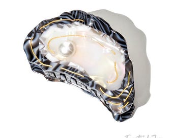 Puede incluir: Una pinza para el pelo con forma de ostra en negro, blanco y dorado. La pinza presenta un adorno de perla y detalles dorados. El diseño imita el aspecto de una concha de ostra, con un interior nacarado y un exterior texturizado.