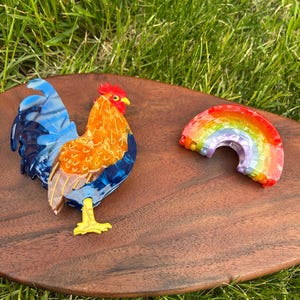 May include: A colorful rooster and rainbow-shaped brooch set on a wooden surface. The rooster features blue, orange, and yellow accents. The rainbow brooch displays a spectrum of colors. The items are set against a backdrop of green grass.