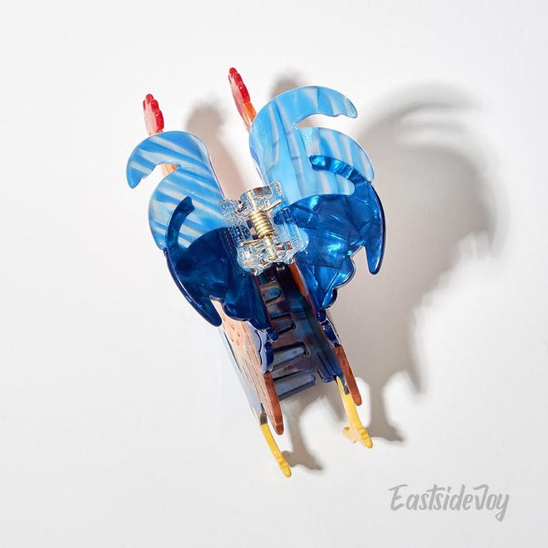 May include: A blue and clear acrylic hair claw clip with a wave design. The clip features a gold-colored spring mechanism and red accents. The clip is approximately 4 inches long.