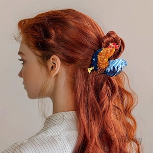 May include: A colorful rooster hair clip, featuring shades of blue, orange, and red, is secured in red wavy hair. The clip is a decorative accessory, adding a whimsical touch to the hairstyle. The background is a neutral color.