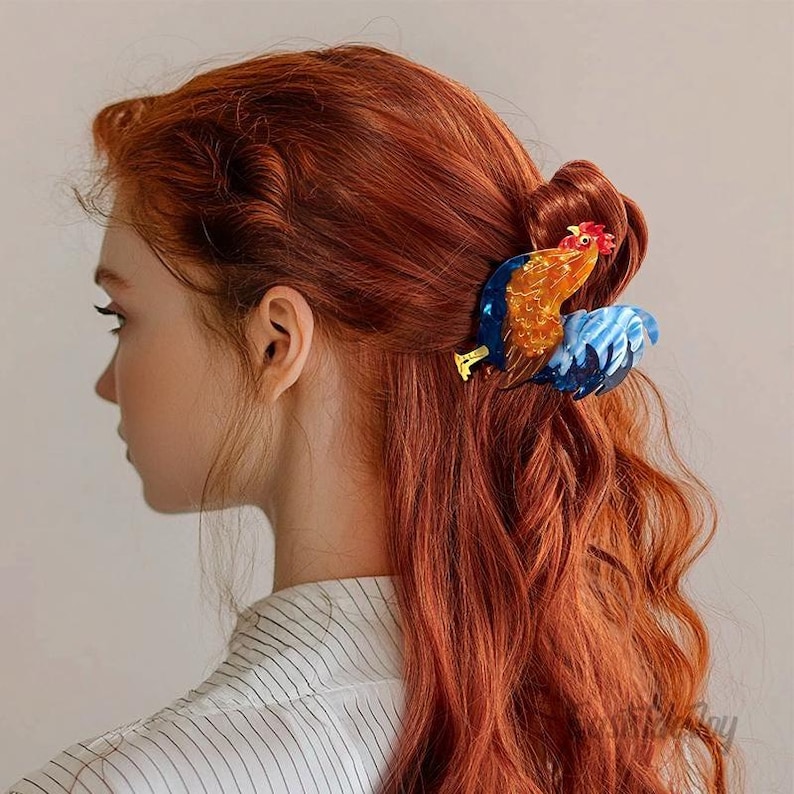 May include: A colorful rooster hair clip, featuring shades of blue, orange, and red, is secured in red wavy hair. The clip is a decorative accessory, adding a whimsical touch to the hairstyle. The background is a neutral color.