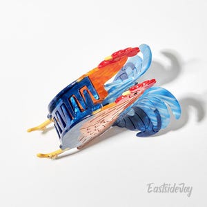 May include: A decorative rooster-shaped hair clip in shades of blue, orange, red, and white. The clip features a detailed design with a rooster's head, body, and tail. The clip is made of a translucent material and has a unique, artistic appearance.