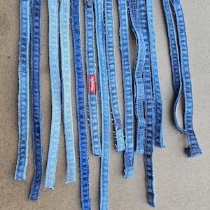 May include: A collection of denim strips in varying shades of blue, arranged on a brown surface. Some strips have a lighter wash, while others are a darker indigo. One strip features a red "Wrangler" label.