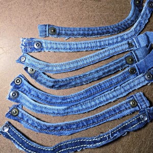 May include: Close-up of several curved denim jean pieces with metal button closures. The pieces are a medium blue wash with visible white stitching and are arranged on a brown surface. The denim has a worn appearance.
