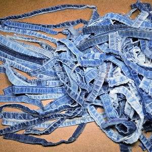 Pode incluir: Uma pilha de tiras de ganga azul, provavelmente reaproveitadas de calças de ganga. As tiras variam em tons de azul, com costuras visíveis e bordas desfiadas. As tiras estão espalhadas sobre uma superfície castanha.