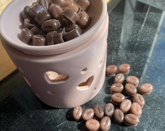 May include: A pink ceramic wax warmer with a heart design, filled with brown coffee bean wax melts.  A pile of additional coffee bean wax melts are scattered on the table.