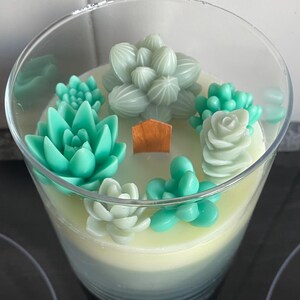 Op de afbeelding: Een heldere glazen potkaars met een gelaagde witte en blauwe wasbasis. De kaars is versierd met groene en lichtgroene was-succulenten.