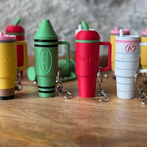 May include: Six colorful, personalized, insulated travel mugs with keychains. The mugs are shaped like a pencil, crayon, apple, and two coffee cups. The mugs are decorated with names and school-themed designs.
