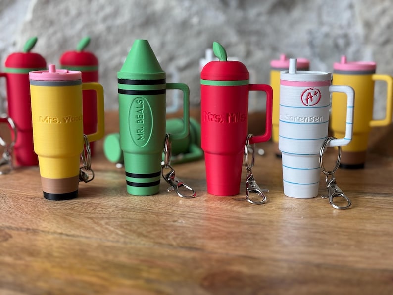 May include: Six colorful, personalized, insulated travel mugs with keychains. The mugs are shaped like a pencil, crayon, apple, and two coffee cups. The mugs are decorated with names and school-themed designs.