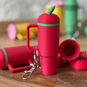 May include: Red silicone keychain with a green leaf on top. The keychain is shaped like a mug with a handle. There are other keychains in the background, including a yellow one and a green one.
