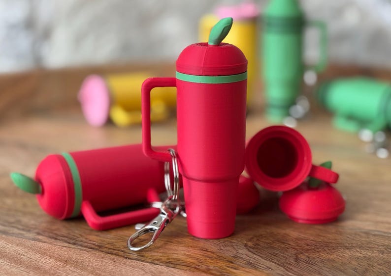 May include: Red silicone keychain with a green leaf on top. The keychain is shaped like a mug with a handle. There are other keychains in the background, including a yellow one and a green one.