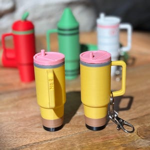 May include: Two yellow pencil-shaped mugs with pink lids and a gray band around the top. The mug on the right has a silver keychain attached to the handle. Both mugs have a brown band around the bottom and a black tip.