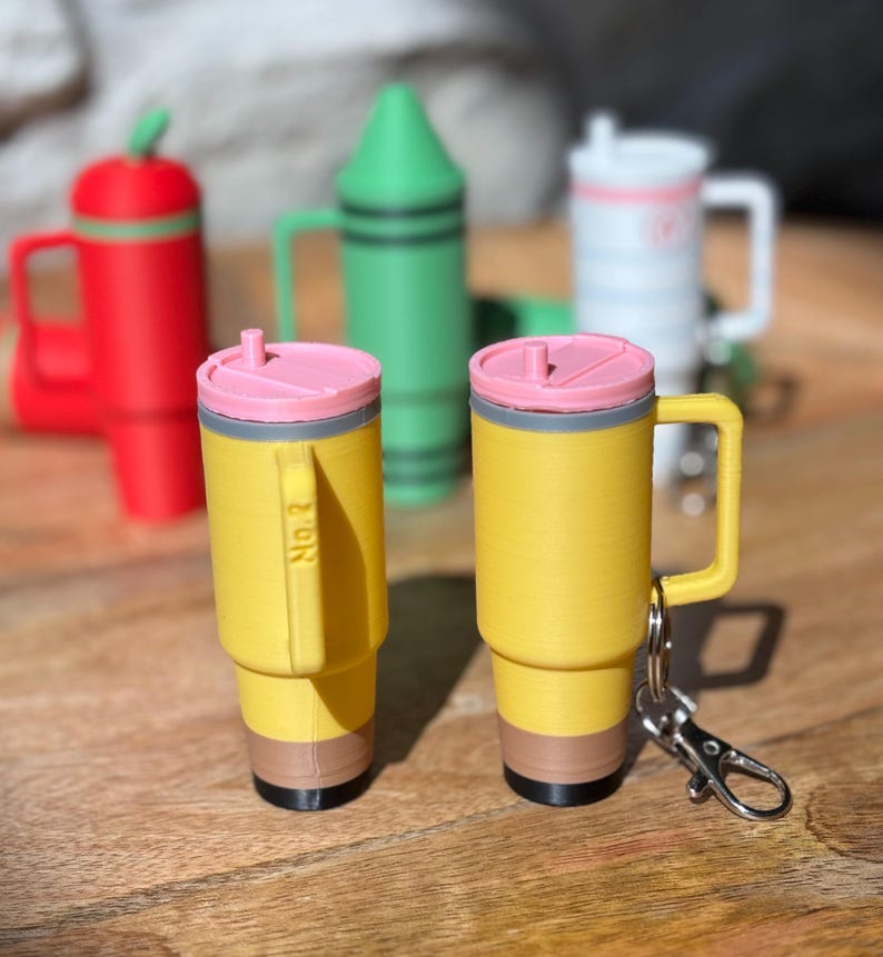 May include: Two yellow pencil-shaped mugs with pink lids and a gray band around the top. The mug on the right has a silver keychain attached to the handle. Both mugs have a brown band around the bottom and a black tip.
