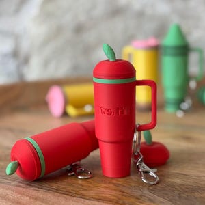 May include: Red apple-shaped keychain with a green leaf on top and a green band around the middle. The keychain has a silver hook and a small red apple charm. The keychain says "Mrs. Miller" on the side.
