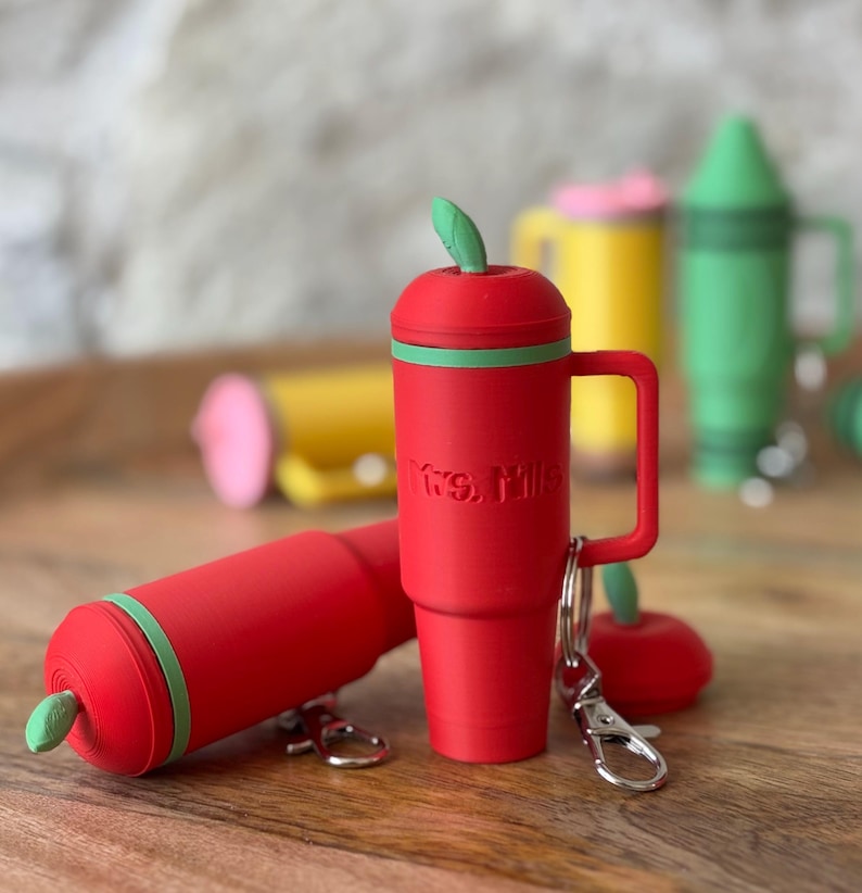 May include: Red apple-shaped keychain with a green leaf on top and a green band around the middle. The keychain has a silver hook and a small red apple charm. The keychain says "Mrs. Miller" on the side.