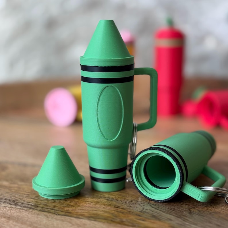 May include: Green crayon-shaped keychain with a black stripe design. The keychain has a small compartment that opens and closes with a lid. The keychain is made of plastic and has a handle.