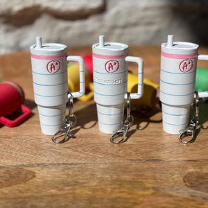 May include: Three white and blue striped cups with pink lids and a red "A+" on the front. Each cup has a silver keychain attached. The cups are sitting on a wooden surface.