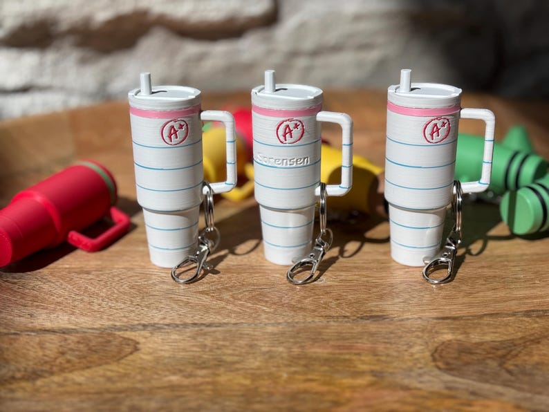 May include: Three white and blue striped cups with pink lids and a red "A+" on the front. Each cup has a silver keychain attached. The cups are sitting on a wooden surface.