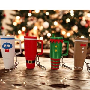 May include: Four insulated travel mugs with handles and keychains. The mugs are white, red, green, and brown, each decorated with a festive design. The white mug has a blue monster face, the red mug has a black belt, the green mug has yellow buttons, and the brown mug has a bow tie.