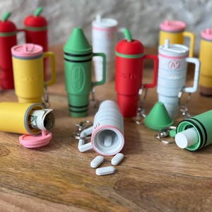 May include: A collection of colorful keychain pill containers in various shapes, including a crayon, a cup, and an apple. Some containers are open, revealing pills and cash inside.