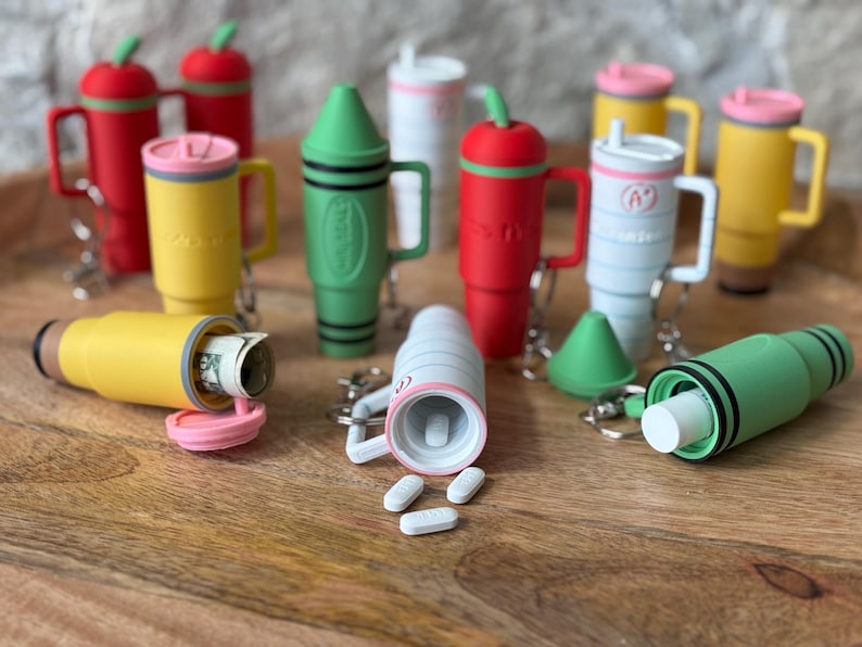 May include: A collection of colorful keychain pill containers in various shapes, including a crayon, a cup, and an apple. Some containers are open, revealing pills and cash inside.