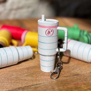 May include: A white and blue notebook-themed keychain with a pink lid and a silver clip. The keychain is shaped like a cup with a handle and a straw. The notebook has a red circle with the text "A+" on it.