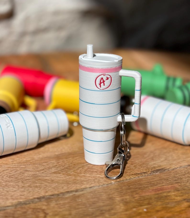 May include: A white and blue notebook-themed keychain with a pink lid and a silver clip. The keychain is shaped like a cup with a handle and a straw. The notebook has a red circle with the text "A+" on it.