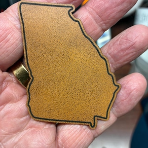 May include: A tan, textured, flat cutout in the shape of the state of Georgia with a thin black outline border. The object has a speckled, grainy surface appearance and is shaped precisely to the geographic borders of the state of Georgia.