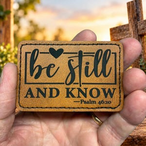 May include: A rectangular tan leather patch with black engraved text that reads be still AND KNOW —Psalm 46:10. The patch features a small black heart icon above the word still and is bordered by a thin black stitched line.