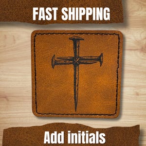 May include: A square, brown leather patch with a stitched border. The patch features a black, stylized cross made of nails. The words "FAST SHIPPING" and "Add initials" are in white text.