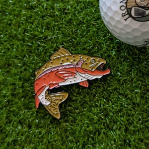 May include: A golf ball with the text "IRON & WOOD GOLF" and a golf club logo, next to a colorful fish-shaped golf ball marker. The marker is brown, red, and gold.
