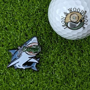 May include: A white golf ball with the text "IRON & WOOD GOLF" and a shark-shaped ball marker. The shark is light blue with a green and white design in its mouth. The items are on a green surface.
