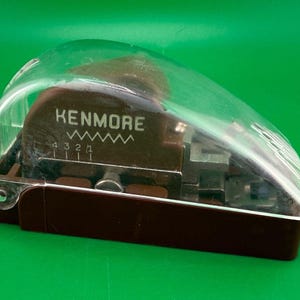 May include: A brown Kenmore sewing machine buttonhole attachment encased in a clear plastic cover. The device features a printed Kenmore logo, a zigzag stitch icon, and a numbered adjustment scale from 1 to 4 on the dark brown body.