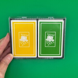 May include: A clear plastic double deck case containing two decks of playing cards. One deck features a yellow back with a white border and a white line drawing of a king. The other deck features a green back with a white border and a white line drawing of a king.
