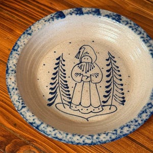 May include: A round ceramic bowl with a speckled off-white base and a blue sponge-painted rim. The interior features a hand-painted blue line drawing of a figure wearing a hooded cloak standing between two stylized evergreen pine trees.
