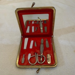 May include: A gold-toned metal manicure kit case with a hinged lid and a rectangular mirror inside. The interior features a bright red molded tray holding various silver-toned metal grooming tools, including scissors, files, and cuticle pushers.