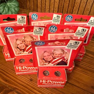 May include: A collection of vintage GE Hi-Power FlashCubes in red and white packaging. Each box features a photograph of a smiling adult and child, the GE logo, and the text GE Flashback Guarantee. The boxes are arranged in a group.