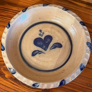 May include: A beige ceramic pie plate with a blue heart design in the center. The plate has a dark blue ring around the heart and a scalloped edge with blue accents. The plate is on a wooden surface.