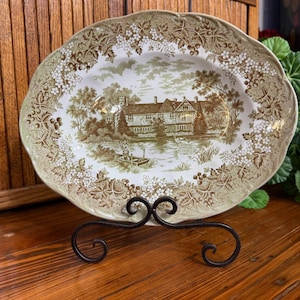 May include: An oval ceramic platter featuring a brown and white transferware pattern of a country manor by a lake with a person in a small boat. The border is decorated with detailed floral and grape vine motifs on a cream-colored background.