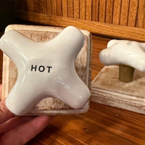 May include: Two vintage-style white ceramic faucet handles. One handle is marked "HOT" in black lettering. The other handle is marked "COLD". Both are mounted on distressed wooden blocks. The handles have a classic, cross-shaped design.