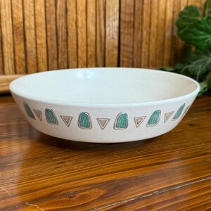 May include: A round, off-white ceramic bowl with a speckled finish. The exterior features a repeating pattern of teal-colored rounded shapes alternating with small, brown-outlined triangles encircling the entire circumference of the bowl.