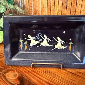 May include: A rectangular dark blue ceramic tray featuring a hand-painted design of three light-colored rabbits leaping across the center. The scene includes small white stars and orange carrots interspersed between the rabbits on the glossy surface.