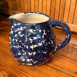 May include: A blue and white speckled ceramic pitcher with a handle and spout. The pitcher is on a wooden surface. The interior of the pitcher is white.