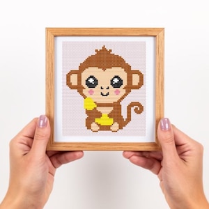 May include: A framed cross-stitch artwork featuring a cartoon monkey holding a banana. The monkey is brown with large, black eyes and a pink mouth. The banana is yellow. The frame is made of light-colored wood.