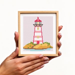May include: A framed cross-stitch of a lighthouse. The lighthouse is pink and white with a red top, set on a small island with green and yellow details. The frame is made of light-colored wood.