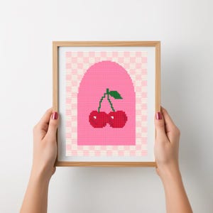 May include: A framed cross-stitch artwork depicting two red cherries with green leaves. The cherries are set against a pink archway on a checkered background, within a wooden frame.
