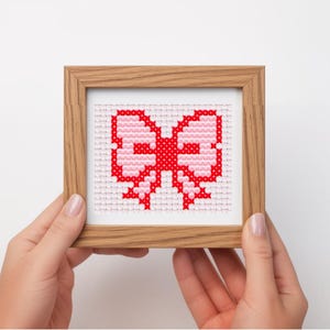 May include: A framed cross-stitch artwork featuring a bow design in shades of red and pink. The bow is centered on a white background, encased in a light brown wooden frame. The artwork is held by two hands.