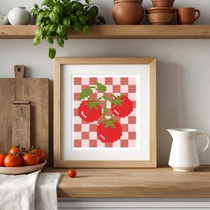 May include: A framed cross-stitch art piece featuring three bright red tomatoes with green stems and leaves, set against a background of a pink and white checkered pattern. The artwork is displayed in a light-colored wooden frame.