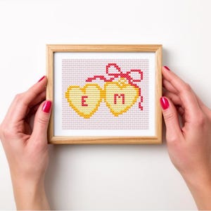 May include: A framed cross-stitch artwork featuring two yellow hearts with the letters "E" and "M" in red. A red bow and ribbon connect the hearts. The artwork is displayed in a light brown wooden frame.